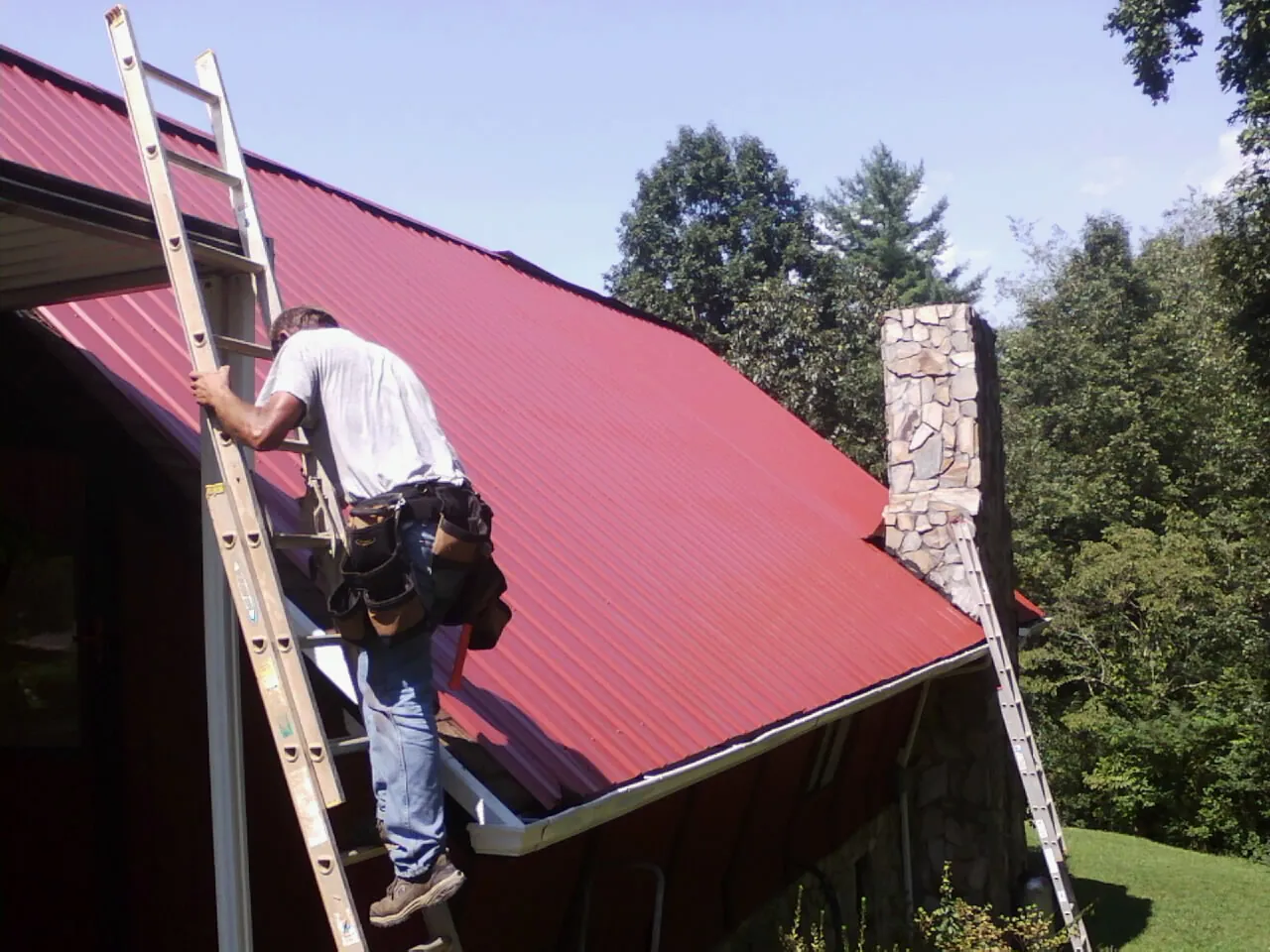 Roofer on a ladder beside a red metal roof and stone chimney — Soffit & Fascia Repair by Roofing Brothers in Elm Mott